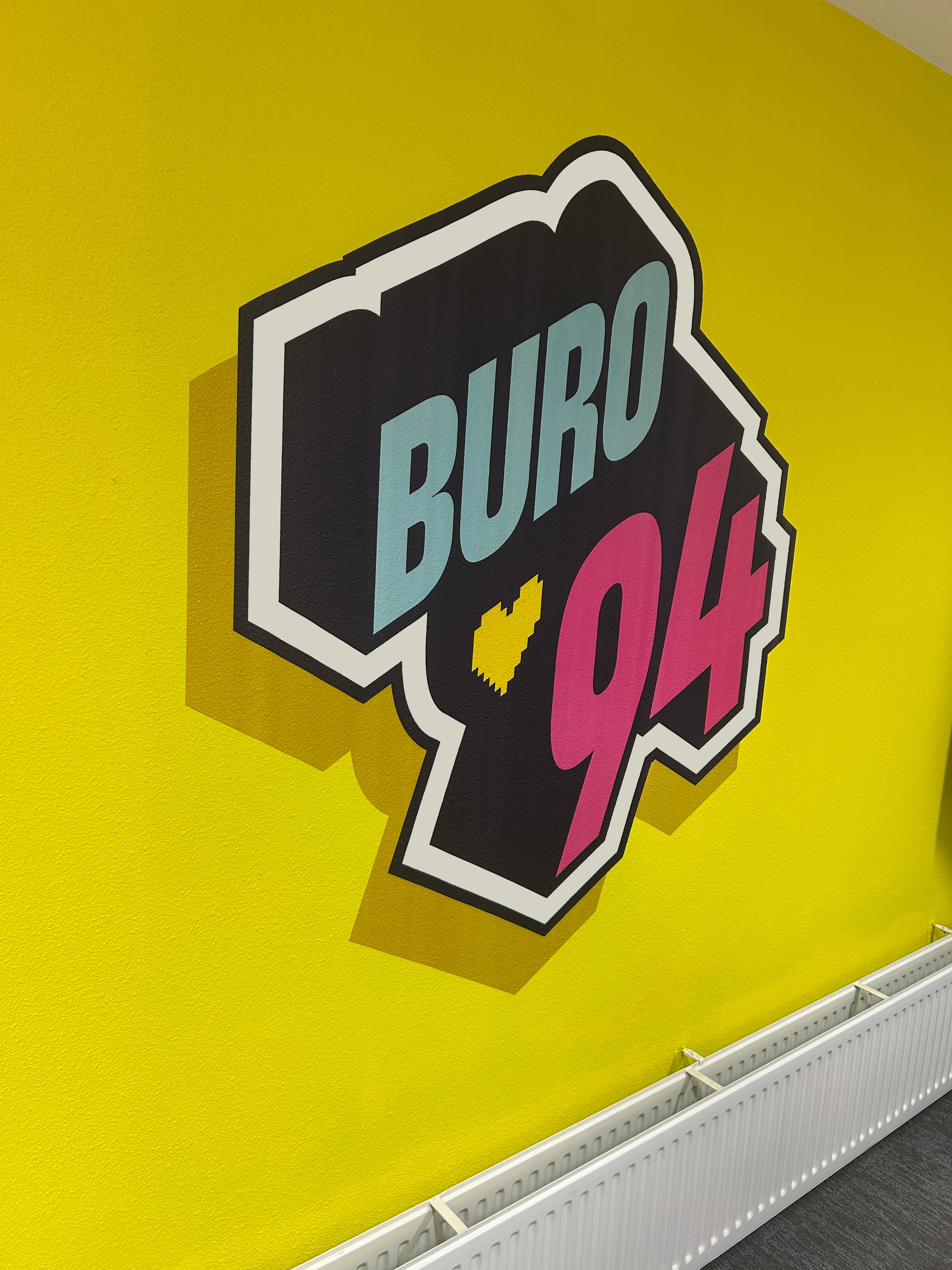 Zakelijke muurprint van het Buro94-logo op een gele spachtelputz kantoorwand, met schaduw en diepte voor een opvallend en strak eindresultaat.